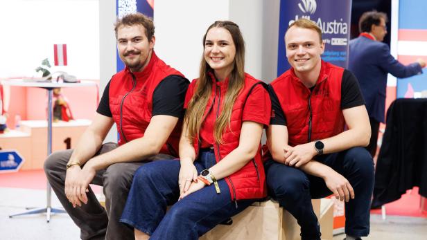 David Kier, Lisa-Marie Spörk und Lukas  Frühwirth sitzen auf Holzboxen vor dem Team-Austria-Stand in Dänemark