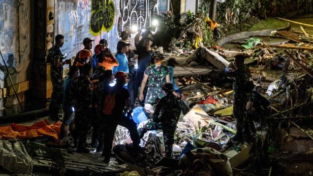 Helfer durchsuchen nachts Trümmer mit Taschenlampen vor einer Wand mit Graffiti.