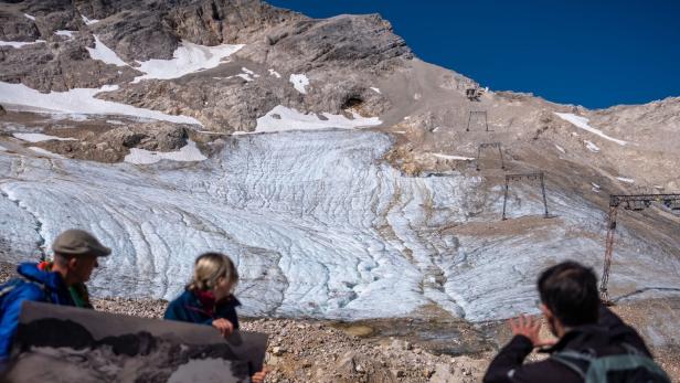Drei Personen stehen vor einem schmelzenden Gletscher in den Bergen, eine Person hält ein altes Foto zum Vergleich.