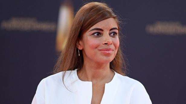 Collien Fernandes mit langen braunen Haaren und weißem Oberteil vor dunklem Hintergrund.