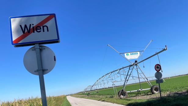 46-218321259 Ein Ortsausgangsschild mit „Wien“ steht an einer Landstraße neben Feldern und einer großen landwirtschaftlichen Bewässerungsanlage.