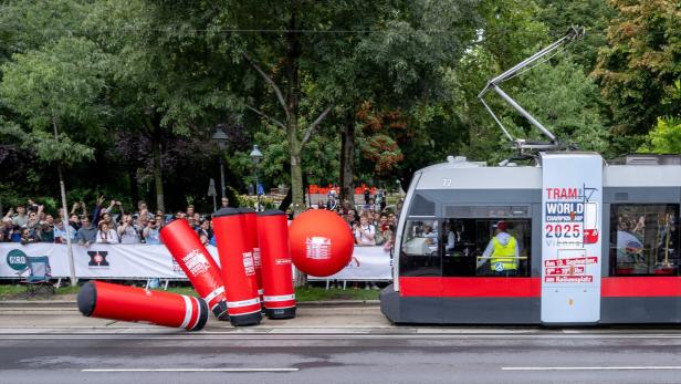 Eine Straßenbahn stößt auf einer Strecke mit großen roten aufblasbaren Hindernissen, während viele Zuschauer am Rand stehen.