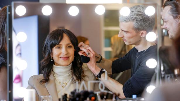 Stephanie Stumph sitzt vor einem beleuchteten Spiegel, während ein Visagist ihr Make-up aufträgt.