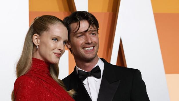 Patrick Schwarzenegger und Abby Champion posieren elegant, sie trägt ein glitzerndes rotes Kleid, er einen schwarzen Smoking mit Fliege, beide lächeln.