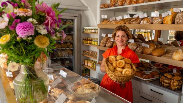 46-218536734 Eine Frau in rotem Kleid hält einen Korb mit Brötchen in einer Bäckerei, umgeben von Broten und einem Blumenstrauß.