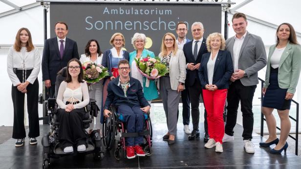 Vierzehn Menschen, darunter zwei im Rollstuhl, stehen und sitzen gemeinsam auf einer Bühne vor einem Schild mit der Aufschrift „Ambulatorium Sonnenschein“.