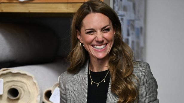 Prinzessin Kate mit langen, gewellten Haaren lacht herzlich und trägt einen karierten Blazer sowie eine goldene Kette mit Anhängern.