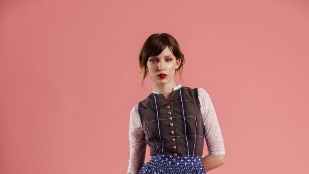 Eine Frau in einem Dirndl mit blauem Rock und karierter Weste vor einem rosa Hintergrund.