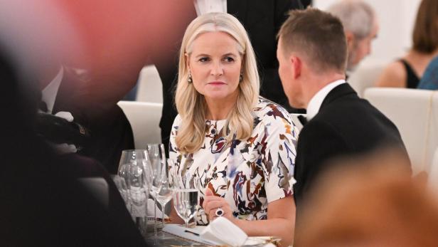 Prinzessin Mette-Marit in einem geblümten Kleid sitzt an einem gedeckten Tisch und spricht mit einem Mann im Anzug.