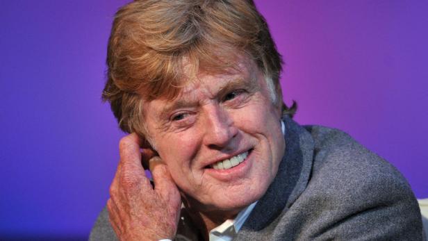 Robert Redford lächelt vor einem violetten Hintergrund und berührt sein Ohr.