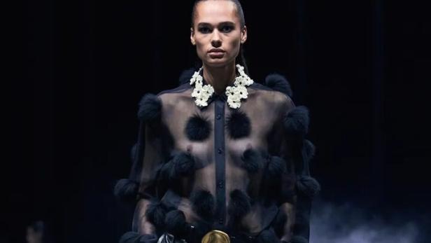 Eine Frau in einer schwarzen, transparenten Bluse mit Fellbommeln und einem schwarzen Ledermantel auf dem Laufsteg.