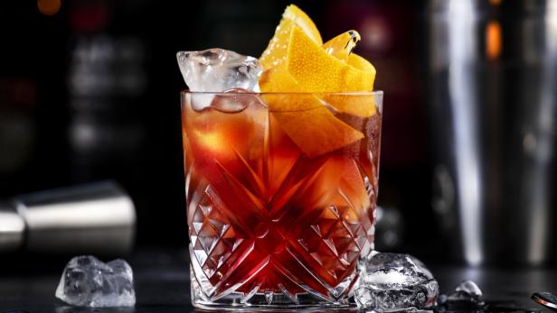 Espresso Negroni alcoholic cocktail drink with dry gin, red vermouth and red bitter, espresso and coffee liqueur, orange slice and ice cubes. Black bar counter background, bar tools, bottles