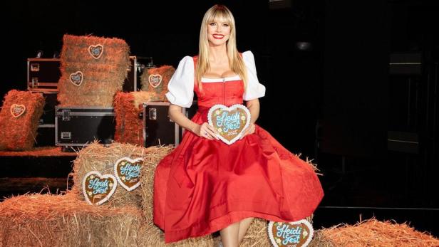 Heidi Klum sitzt im Dirndl mit Lebkuchenherz in Händen auf einem Strohballen