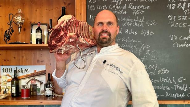 Der Wirt Hannes Tschida trägt ein großes Stück Fleisch auf der Schulter. Auf der Tafel hinter ihm ist die Weinkarte mit Kreide aufgemalt.  