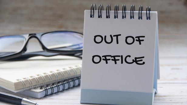 Ein Tischkalender zeigt die Aufschrift „OUT OF OFFICE“, daneben liegen eine Brille, Notizbücher und ein Stift.