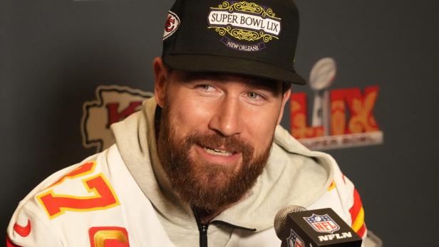 Travis Kelce.jpg Travis Kelce mit Bart und einer "Super Bowl LIX"-Kappe steht vor einem Mikrofon.