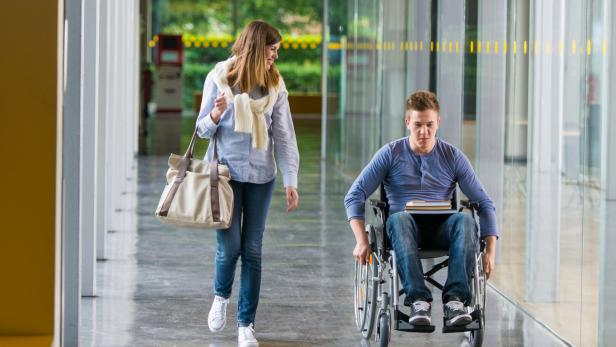 Eine junge Frau geht neben einem jungen Mann im Rollstuhl einen Flur entlang.