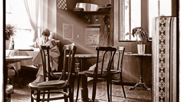 Eine Frau sitzt im Kaffeehaus am Fenster, Sonnenlicht fällt rein. Sie liest Zeitung.