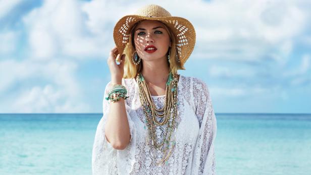 Eine Frau mit Hut, Schmuck und weißer Spitze vor einem Strand.