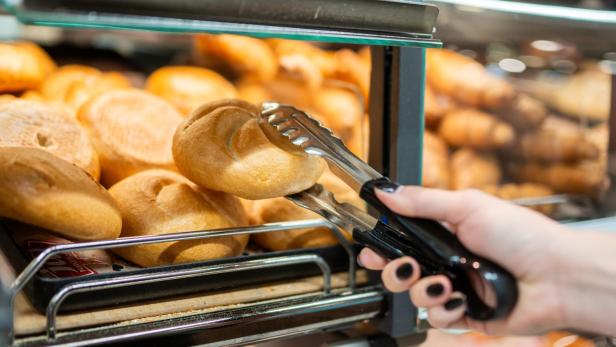 Eine Hand nimmt mit einer Zange ein Brötchen aus einer Auslage.