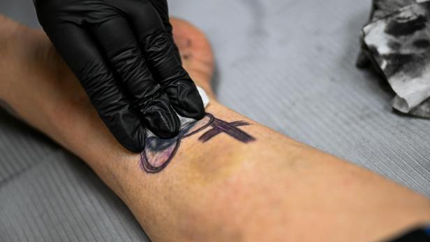 Eine Hand mit schwarzem Handschuh reinigt ein frisches Tattoo am Bein mit einem Tuch.
