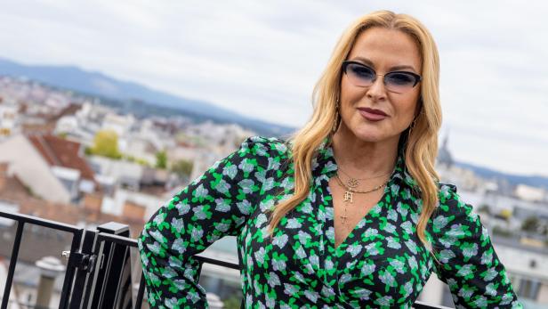 Anastacia mit Sonnenbrille und blumigem Kleid posiert vor einer Stadtkulisse.