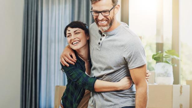 Ein Mann und eine Frau befinden sich in einer Wohnung mit Umzugskartons im Hintergrund und umarmen sich lächelnd.