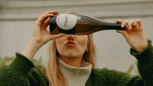 Winzerin Katharina Gessl steht zwischen ihren Weinstöcken, hält eine ihrer Weinflaschen vors Gesicht und zeigt das Etikett. 