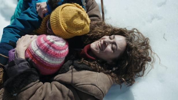Eine Frau mit langen Haaren liegt im Schnee und umarmt ihre zwei Kinder.