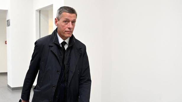 Ein Mann in einem dunklen Mantel geht mit einer Aktentasche einen Flur entlang.