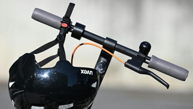 Ein schwarzer Fahrradhelm hängt am Lenker eines E-Scooters mit grauen Griffen und orangefarbenem Kabel.