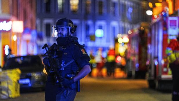 Ein Polizist mit Helm und Gewehr steht auf einer belebten Straße bei Nacht.
