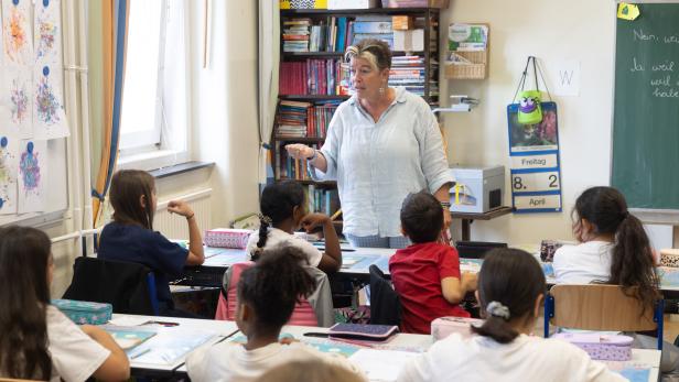 Eine Lehrerin spricht vor einer Gruppe von Grundschulkindern, die an ihren Tischen sitzen und zuhören.