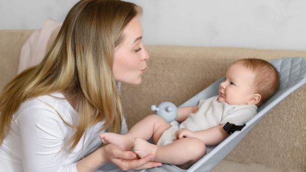 Eine Frau beugt sich lächelnd zu einem Baby, das entspannt in einer Wippe liegt.