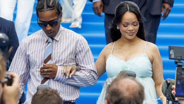 Rihanna und A$AP Rocky schreiten Hand in Hand vor blauem Hintergrund an Fotografen vorbei.
