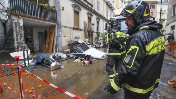 Zwei Feuerwehrleute stehen vor einem Haus, dessen Eingang von Schlamm und Trümmern nach einer Überschwemmung blockiert ist.