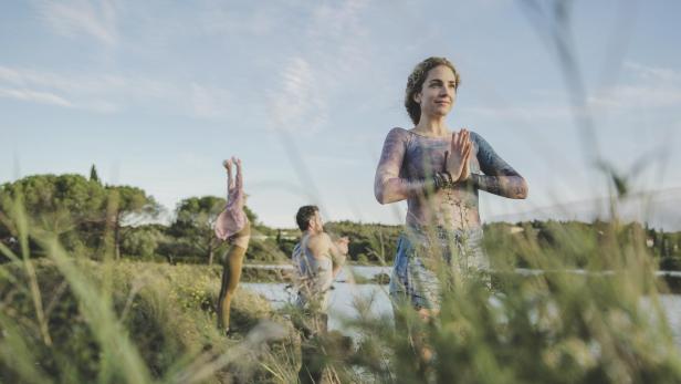 Drei Personen machen Yoga im Grünen, im Hintergrund das Meer.