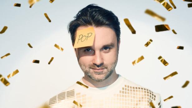 Ein Mann - Dr. Pop - mit Mikrofon und einem Zettel mit der Aufschrift „POP“ auf der Stirn steht im goldenen Konfettiregen.