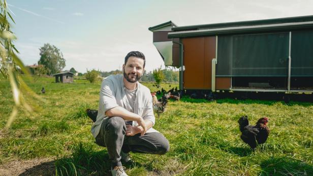 Ein Mann hockt auf einer Wiese mit Hühnern vor einem mobilen Hühnerstall.