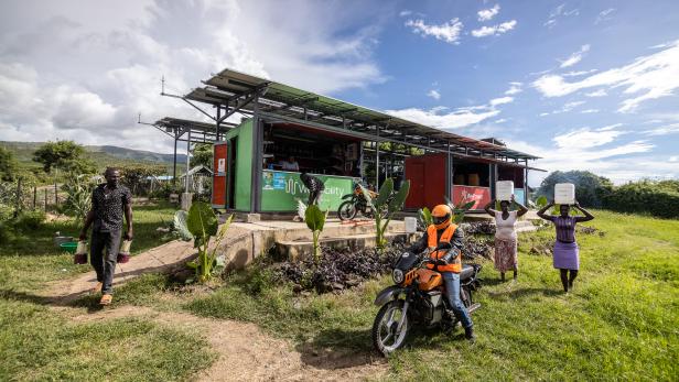 Grünes Containergebäude mit Solarpanelen auf dem Dach, davor eine Person auf einem Motorrad mit orangefarbenem Helm und Weste, zwei Frauen tragen Behälter auf dem Kopf, ländliche Umgebung mit Gras und Bäumen.