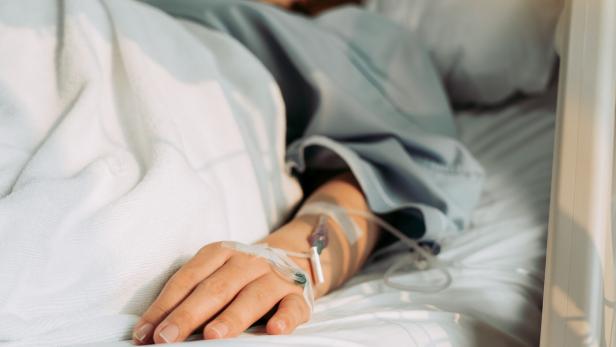 Eine Person liegt im Krankenhausbett, ein Infusionsschlauch ist an ihrem Arm befestigt.