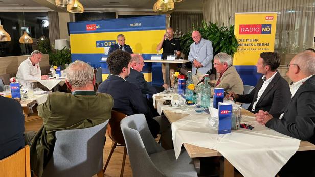 Mehrere Männer sitzen in einem Wirtshaus, im Hintergrund sind Banner des ORF-Niederösterreich zu sehen und Martin Gebhart spricht in ein Mikrofon.
