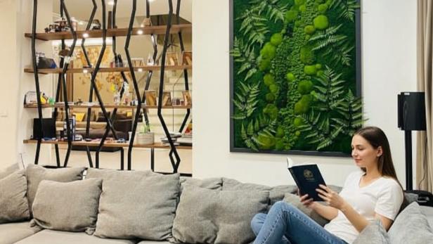 Eine Frau sitzt im Wohnzimmer auf der Couch und liest ein Buch. Im Hintergrund sieht man ein großes Moosbild an der Wand, das eine gemütliche Atmosphäre schafft.