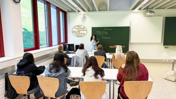 Mehrere Schülerinnen und Schüler sitzen in einem Klassenraum und hören einer Lehrerin zu, die an der Tafel steht und erklärt.