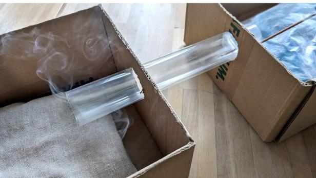 Zwei Kartons sind mit einem durchsichtigen Rohr verbunden, wobei aus einem Karton Rauch aufsteigt.