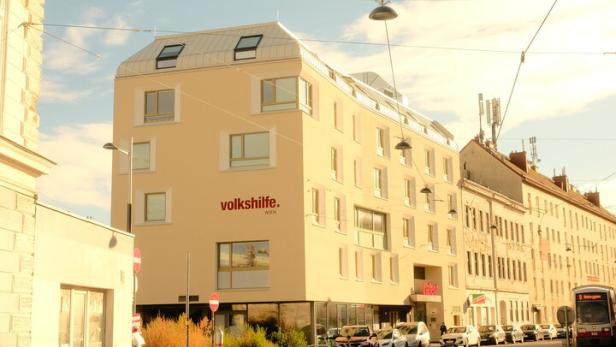 Mehrstöckiges, modernes Gebäude mit der Aufschrift „Volkshilfe“ an einer Straßenecke, mehrere geparkte Autos und Straßenbahn.