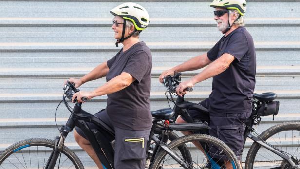 Happy senior couple doing together outdoor activity with electric bicycle. Healthy lifestyle. Yellow helmet, black clothes. Outdoor against a gray metal panel