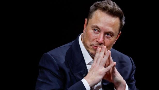 Elon Musk in Anzug sitzt nachdenklich mit verschränkten Händen vor schwarzem Hintergrund.
