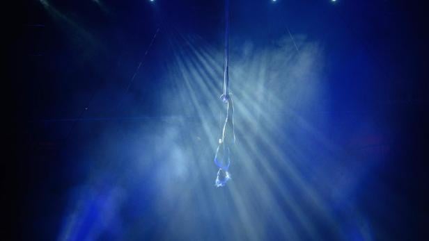 Eine Artistin hängt kopfüber an einem Seil im blauen Scheinwerferlicht.