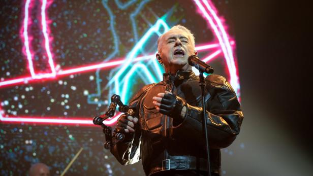 Ein Sänger mit grauen Haaren - Holly Johnson - performt in Lederjacke vor einer Bühne mit Neonlichteffekten.